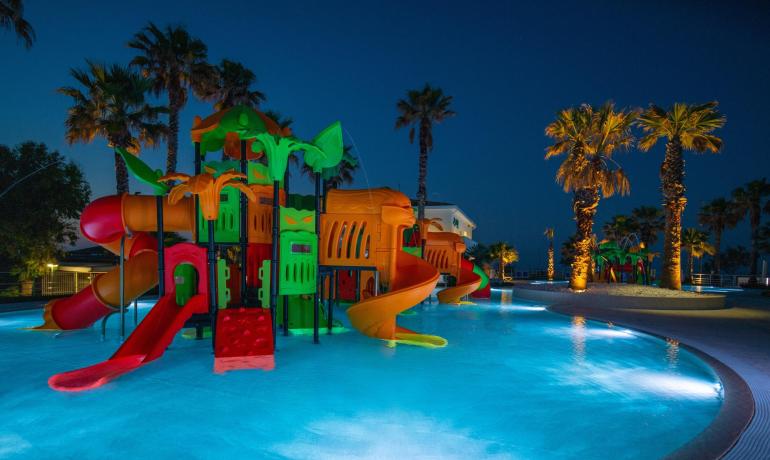 Parco giochi acquatico illuminato di notte, con scivoli colorati e palme.